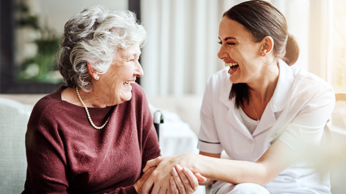 Eine ältere Frau und eine junge Pflegerin halten sich an den Händen und lachen herzlich
